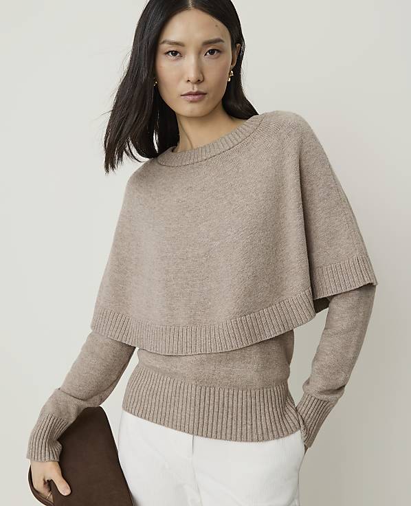 A woman in a beige sweater with a cape overlay stands against a plain, light background.