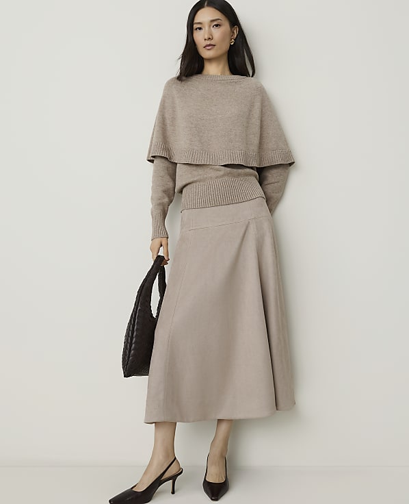 A woman in a beige sweater and skirt stands against a plain white background, holding a dark brown handbag.