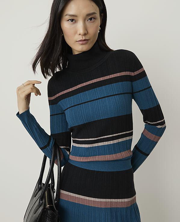 A woman in a striped turtleneck sweater and skirt stands against a plain background, holding a black handbag.