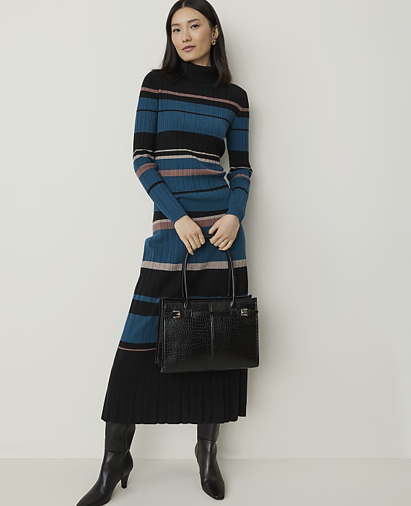 A woman in a striped turtleneck dress and black boots holds a black crocodile-embossed handbag against a plain white background.