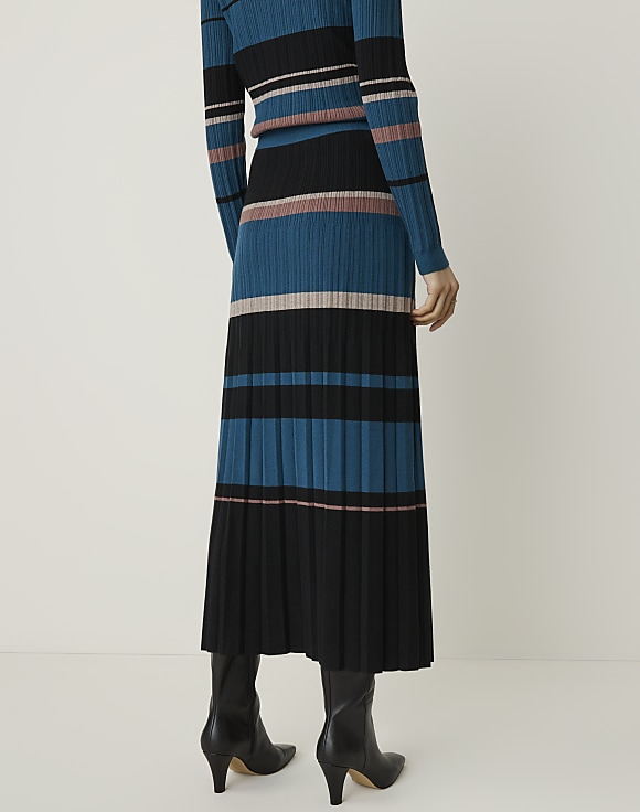 A person stands in black boots, wearing a pleated midi skirt and a matching long-sleeved top with horizontal stripes of teal, black, and pink against a plain, light background.