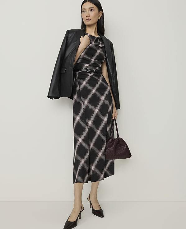 A woman in a plaid dress and leather jacket stands against a plain background, holding a dark handbag.