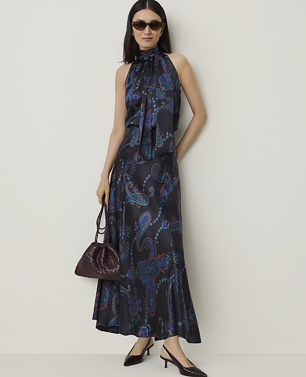 A woman in a patterned halter dress and sunglasses stands with a handbag in the foreground against a plain wall in the background.