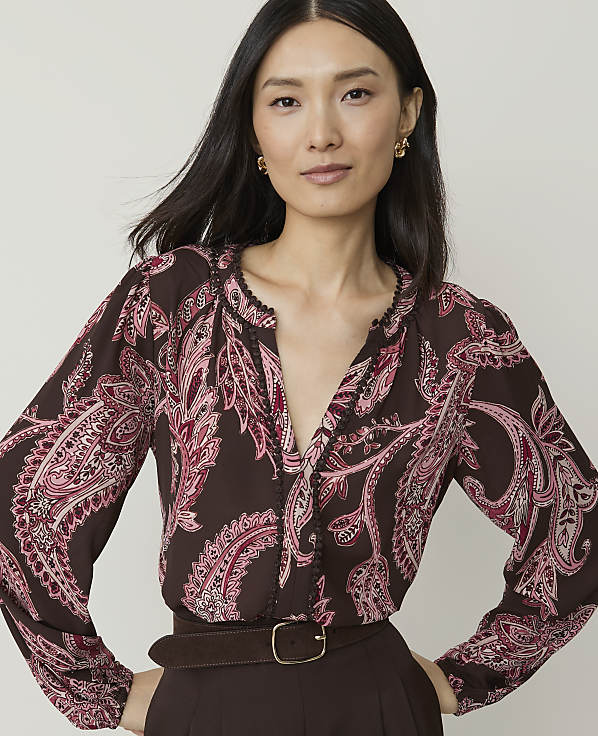 A woman with dark hair and a patterned blouse stands against a plain background.