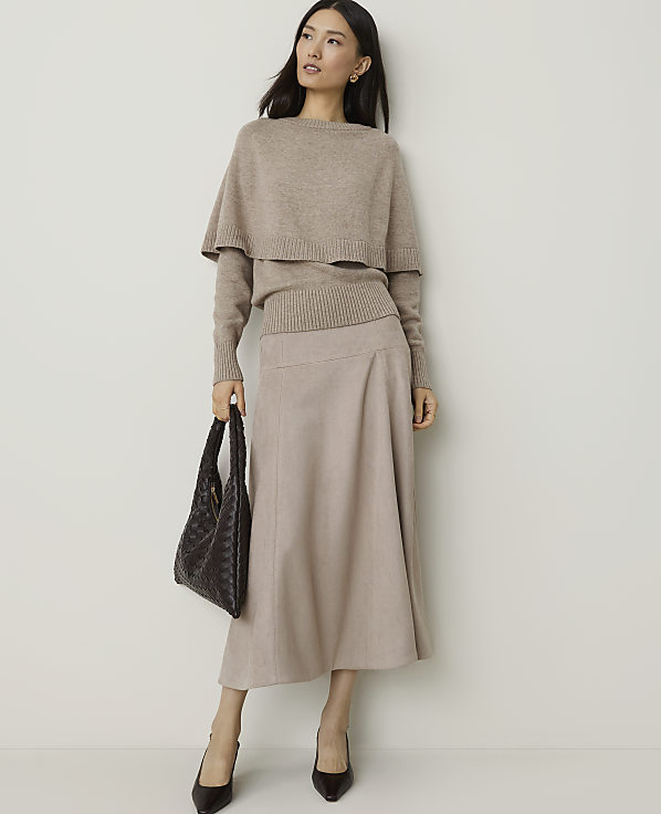 A woman in a beige sweater and skirt stands against a plain white background, holding a dark woven handbag.
