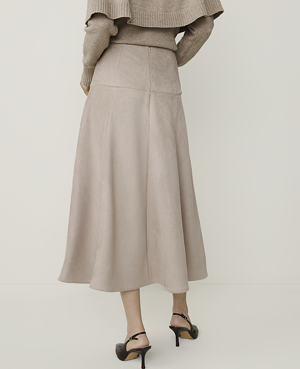 A woman in a beige sweater and a flowing beige skirt stands against a plain white background.