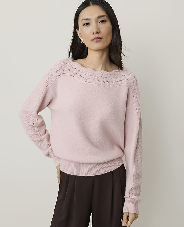 A woman in a pink sweater and brown pants stands against a plain background.