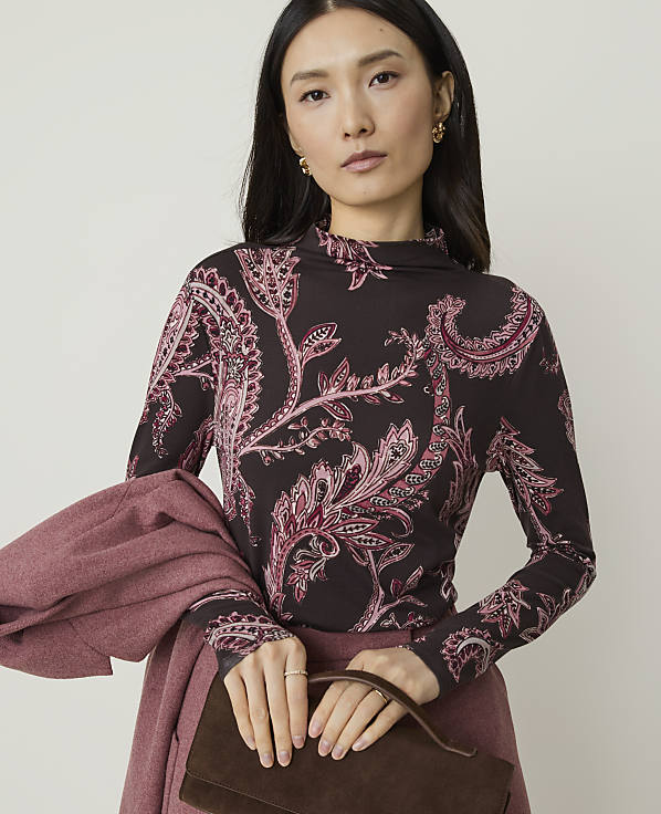 A woman in a patterned top and pink skirt holds a brown clutch in front of a plain background.
