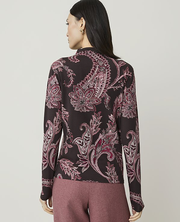 A woman in a dark, paisley-patterned top and pink pants stands against a plain, light gray background.