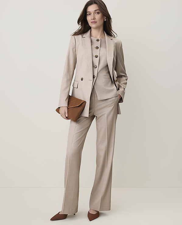 A woman in a beige pantsuit and vest holds a brown clutch in front of a plain, light-colored background.