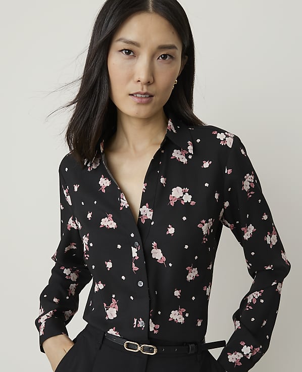 A woman with dark hair and fair skin wears a black floral-print blouse and black pants, with a plain background.