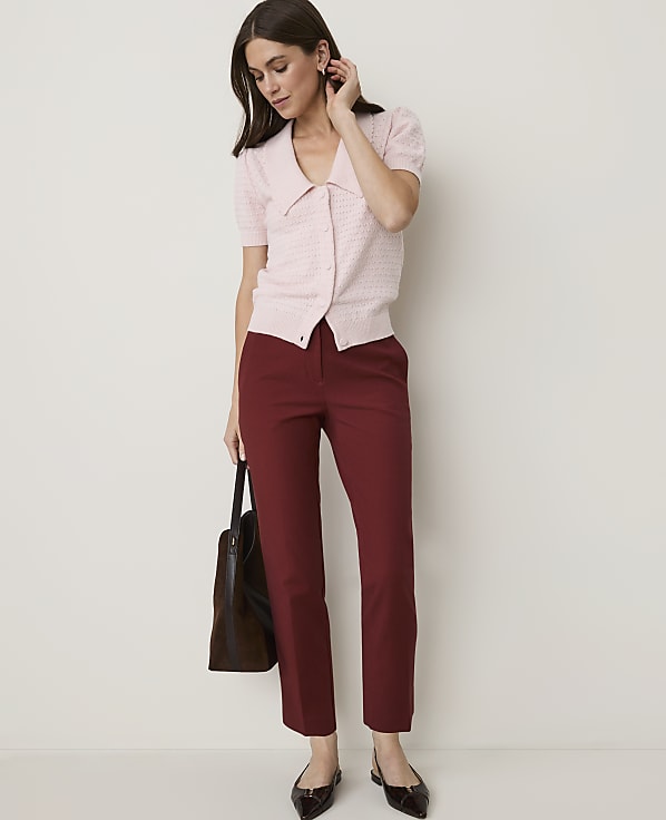 A woman in a pink cardigan and maroon pants stands against a plain white background, holding a dark tote bag.