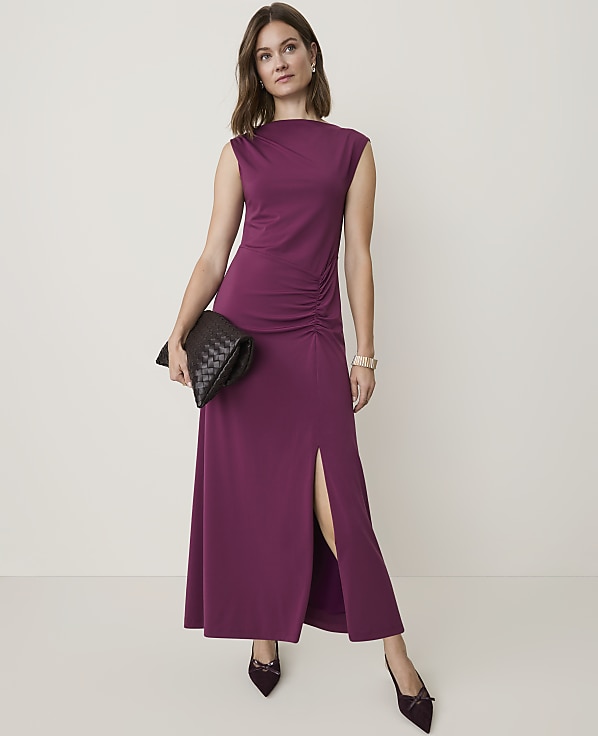 A woman in a plum-colored maxi dress with a high neckline and ruched detailing stands against a plain, light background.