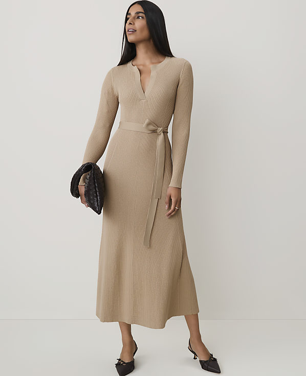A woman in a beige wrap dress and heels holds a dark clutch in front of a plain white background.