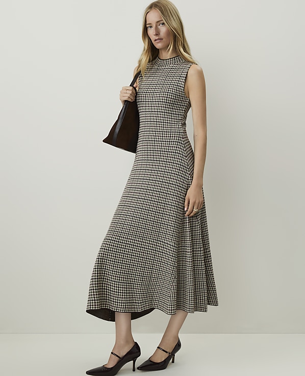 A woman in a patterned dress and heels stands with a bag over her shoulder against a plain background.