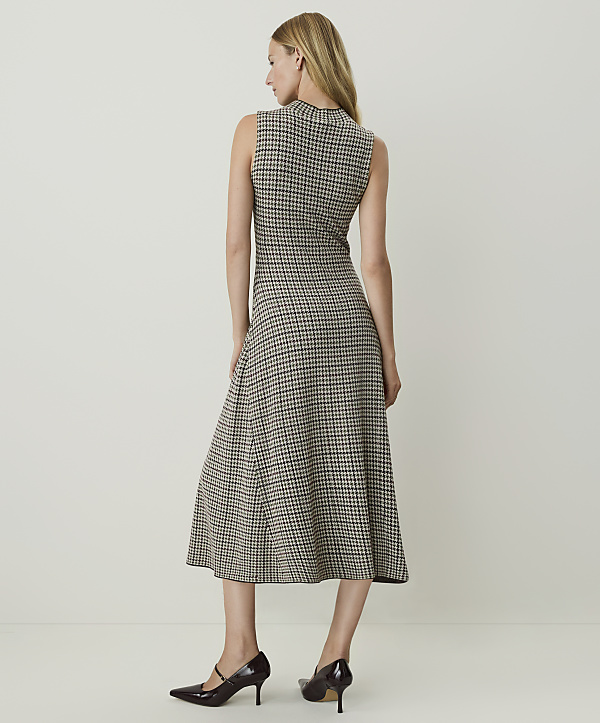 A woman in a sleeveless houndstooth dress and dark heels stands against a plain, light-colored background.