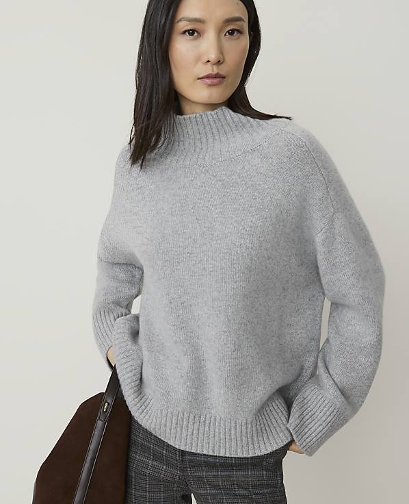 A woman in a grey sweater and plaid pants stands against a plain background, with a brown bag on her left side.