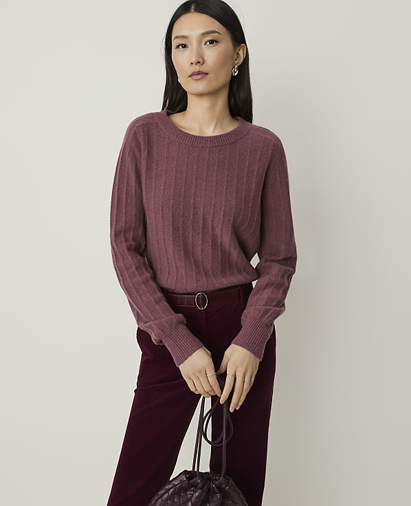 A woman in a mauve ribbed sweater and burgundy pants holds a quilted drawstring bag against a plain, light gray background.
