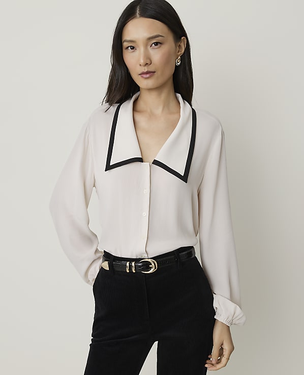 A woman in a cream blouse with a black collar and black pants stands against a plain, light background.