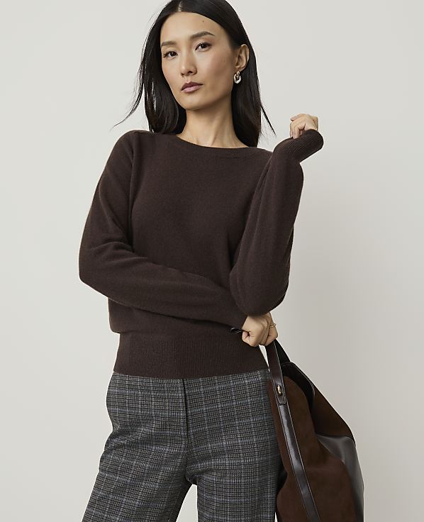 A woman in a brown sweater and plaid pants stands against a plain background, holding a brown bag.