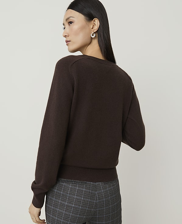 A woman in a brown sweater and plaid pants stands against a plain background.