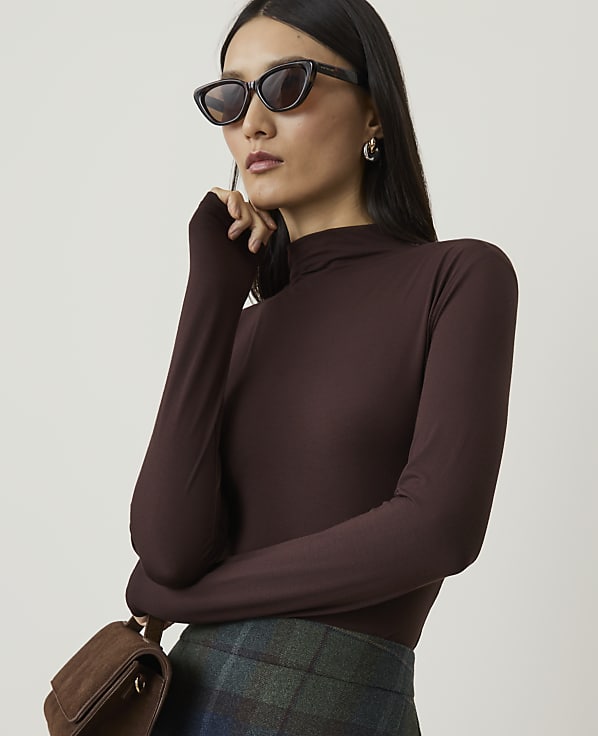 A woman in a brown turtleneck and sunglasses poses with a brown purse in front of a plain background.