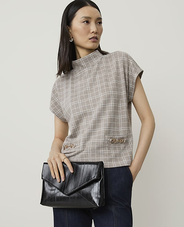 A woman in a plaid top and dark pants holds a black clutch in front of a plain background.