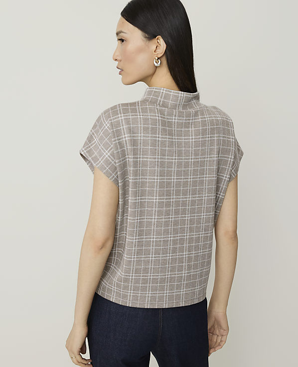 A woman in a plaid top and dark pants stands against a plain, light-colored background.