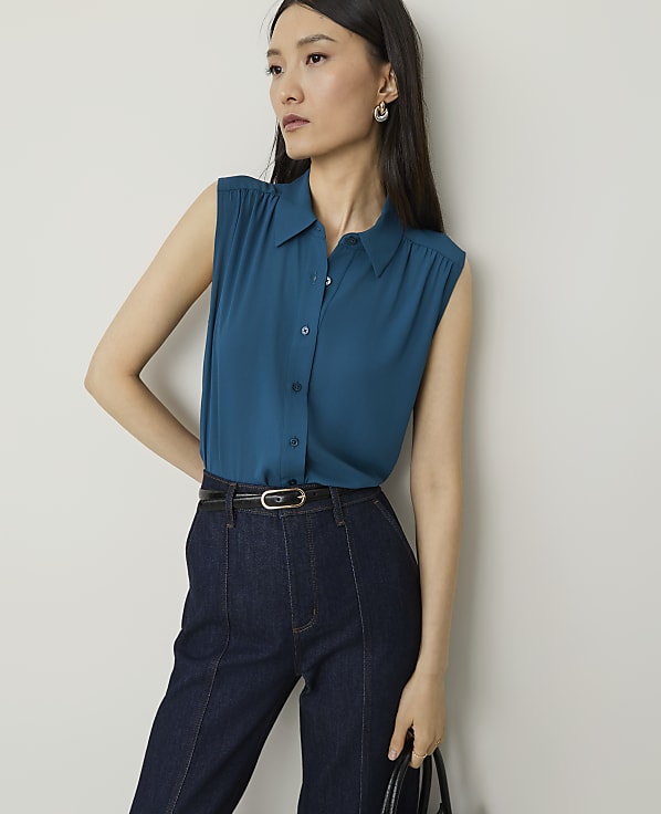 A woman in a teal sleeveless blouse and dark wash jeans stands against a plain white background.