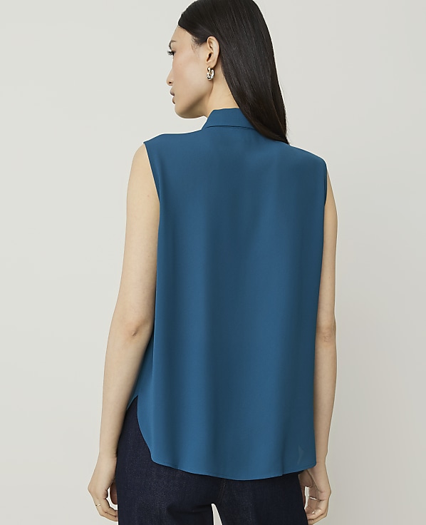 A woman in a teal sleeveless top and dark jeans stands against a plain, light gray background.