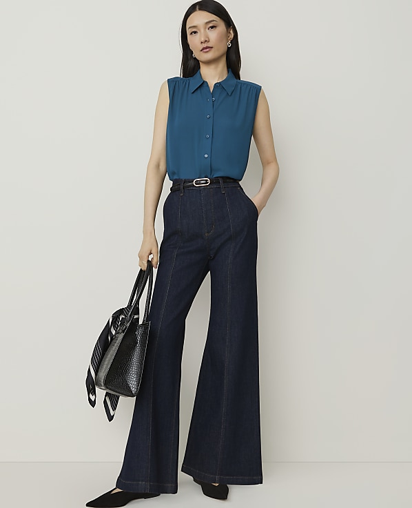 A woman in a teal sleeveless blouse and dark wash wide-leg jeans stands against a plain white background, holding a black tote bag.