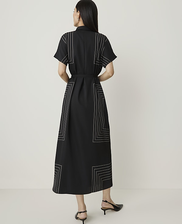 A woman in a black midi dress with geometric stitching stands against a plain, light-colored background.