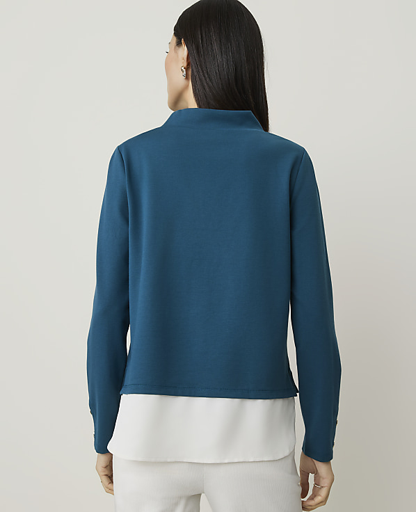 A woman in a teal mock-neck top and white pants stands against a plain, light background.