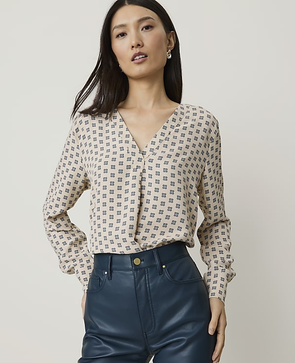 A woman in a patterned blouse and teal leather pants stands against a plain background.