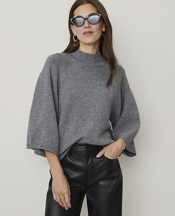 A woman in a grey sweater and black leather pants poses against a plain background.