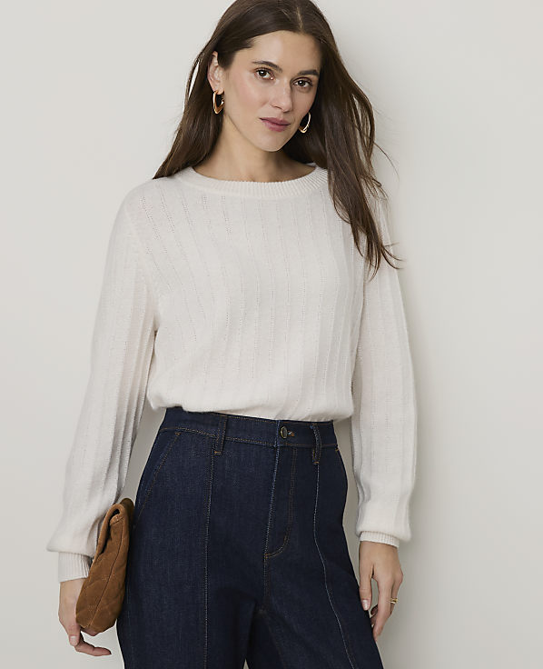 A woman in a cream sweater and dark jeans stands against a plain, light-colored background, holding a brown clutch.