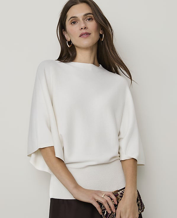 A woman in a cream-colored sweater and dark skirt poses against a plain, light background.