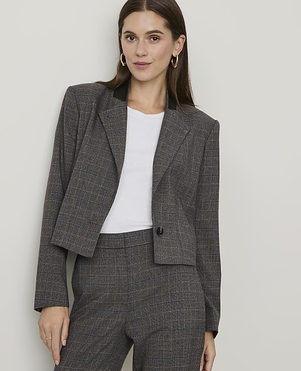 A woman in a plaid blazer and pants stands against a plain, light-colored wall.