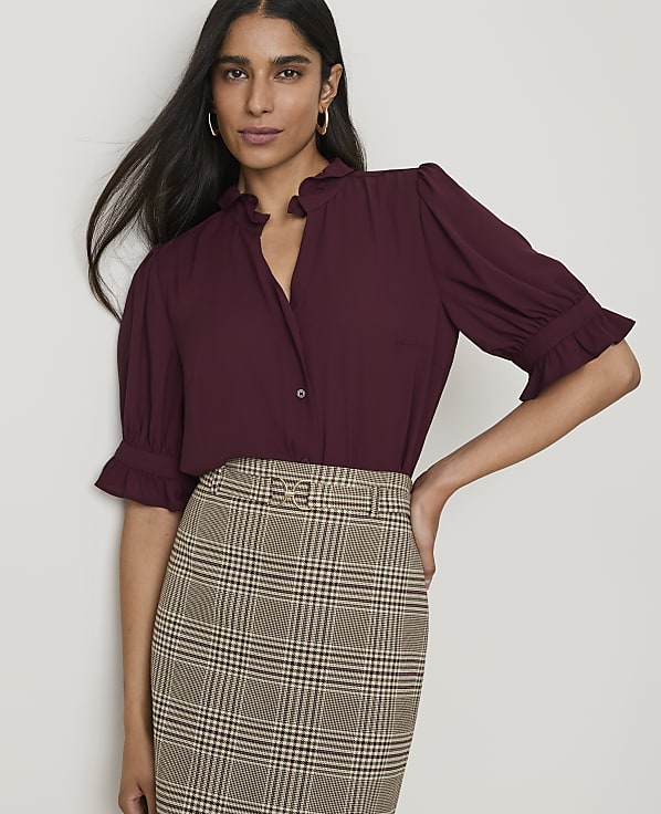 A woman in a burgundy ruffled blouse and plaid skirt stands against a plain white background.
