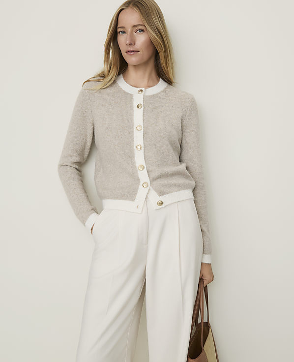 A woman in a beige cardigan and white pants stands against a plain, light-colored background, holding a brown tote bag.
