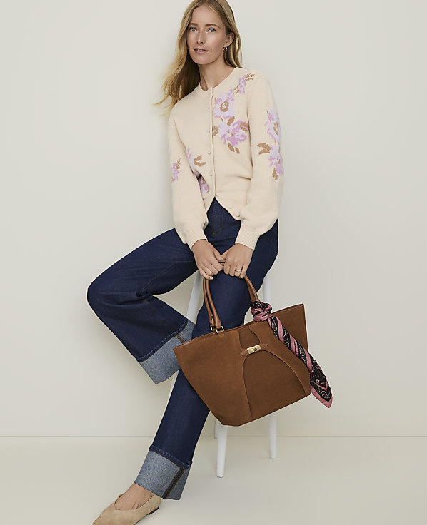 A woman in a cream floral cardigan and dark wash jeans sits on a stool holding a brown suede tote bag, with a plain white background.