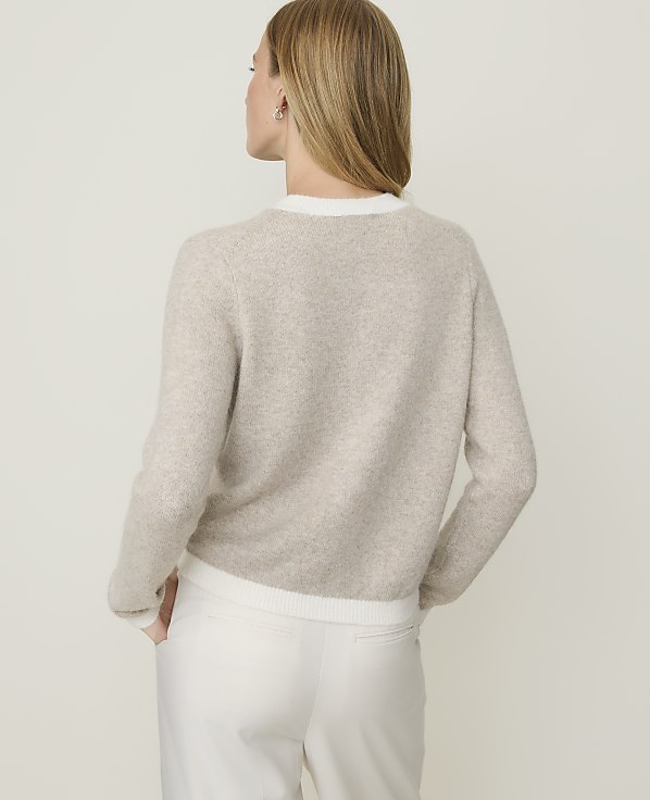 A woman in a beige sweater and white pants stands against a plain, light background.
