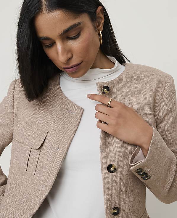 A woman in a beige tweed jacket and white turtleneck stands against a plain, light background.