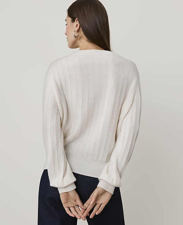A woman in a cream-colored sweater and dark pants stands with her back to the camera against a plain white background.