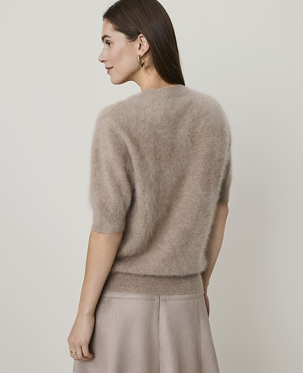 A woman in a fuzzy brown sweater and a beige skirt stands against a plain, light brown background.