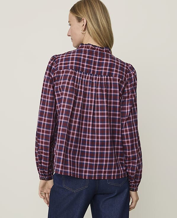A woman in a maroon and pink plaid blouse stands against a plain, light-colored background.