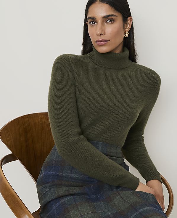 A woman in a green turtleneck and plaid skirt sits on a wooden chair against a plain background.
