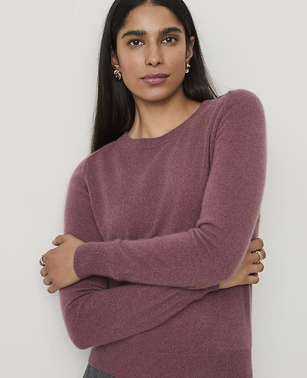 A woman in a mauve sweater crosses her arms in front of a plain white background.