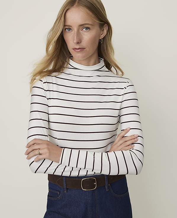 A woman with blonde hair and fair skin, wearing a striped turtleneck and dark jeans, stands against a plain, light-colored background.