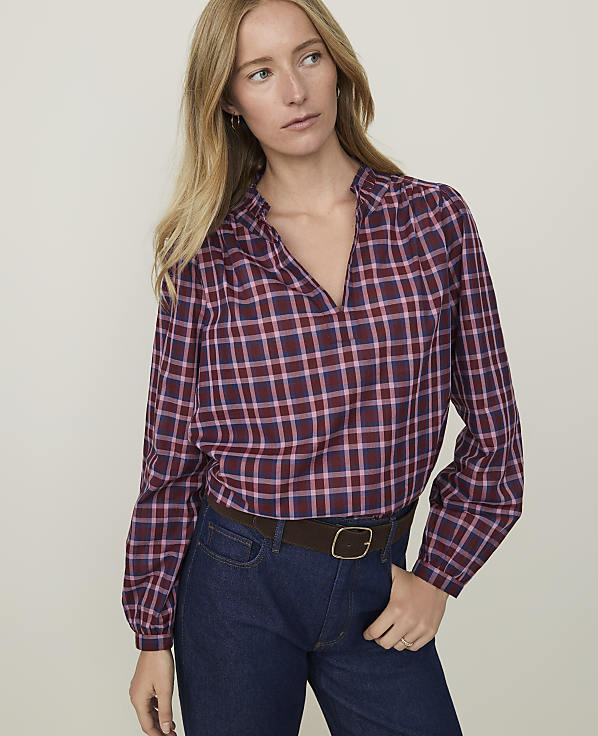 A woman in a maroon and blue plaid blouse and dark jeans stands against a plain, light background.
